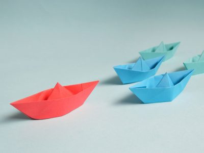 a red origami boat leading the way for several other blue and green origami boats