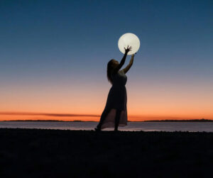 woman on a beach at sunset holding up the moon